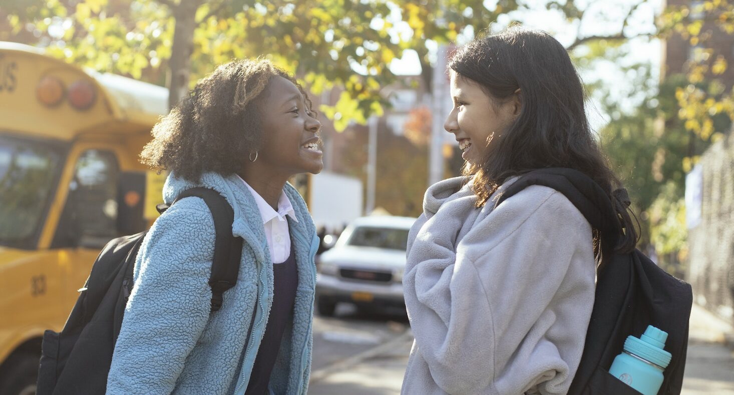 Side view happy multiracial schoolgirls in warm jackets with backpacks standing on street near school bus and talking while looking at each other with smile