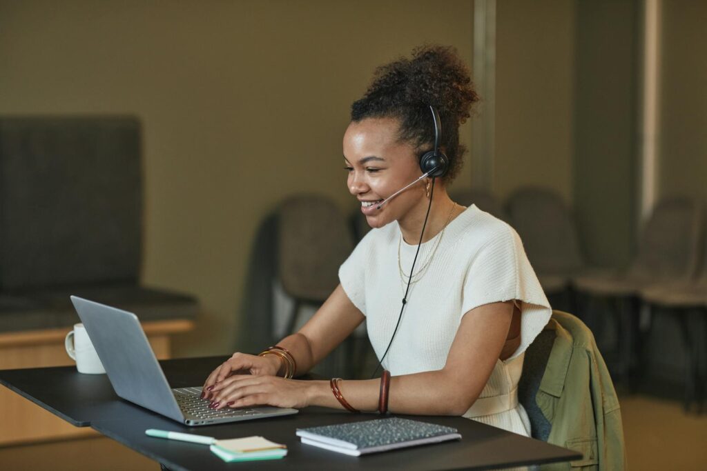 A Woman with Curly Hair Working while Smiling. She is wearing a headset with a microphone placed next to her mouth. This allows for better audio clarity both in terms of hearing her client, but also having the client hear her instructions. 