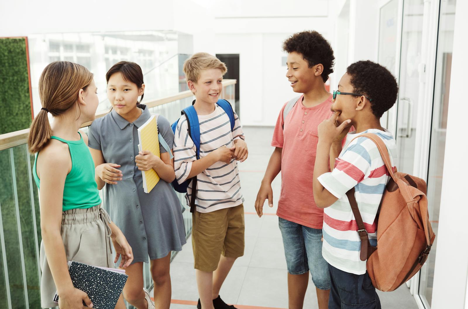 Diverse group of students talking and laughing in a school hallway, holding books and bags.